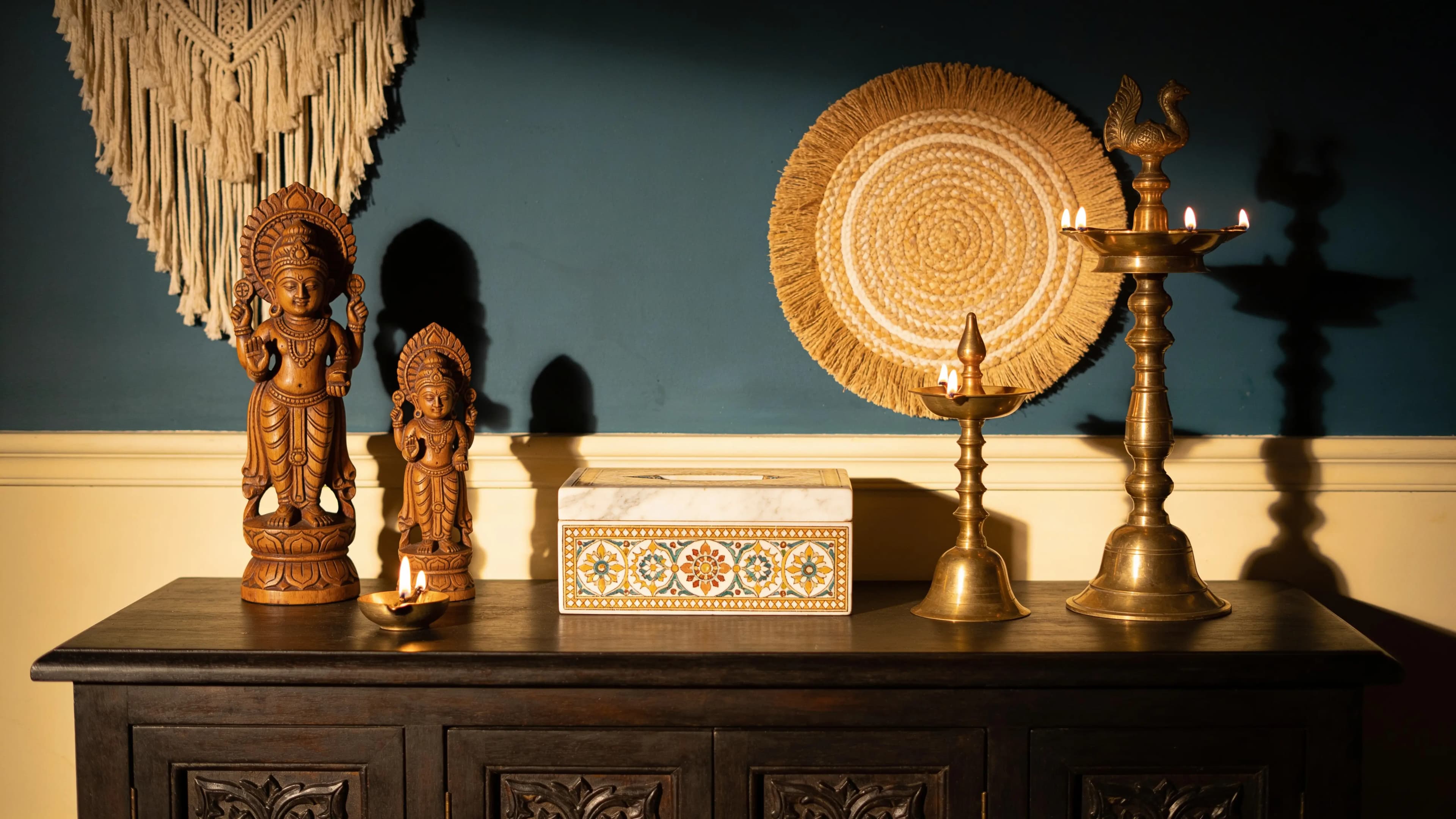 Carved wooden figures, brass lamps and a woven wall plate on a dark sideboard